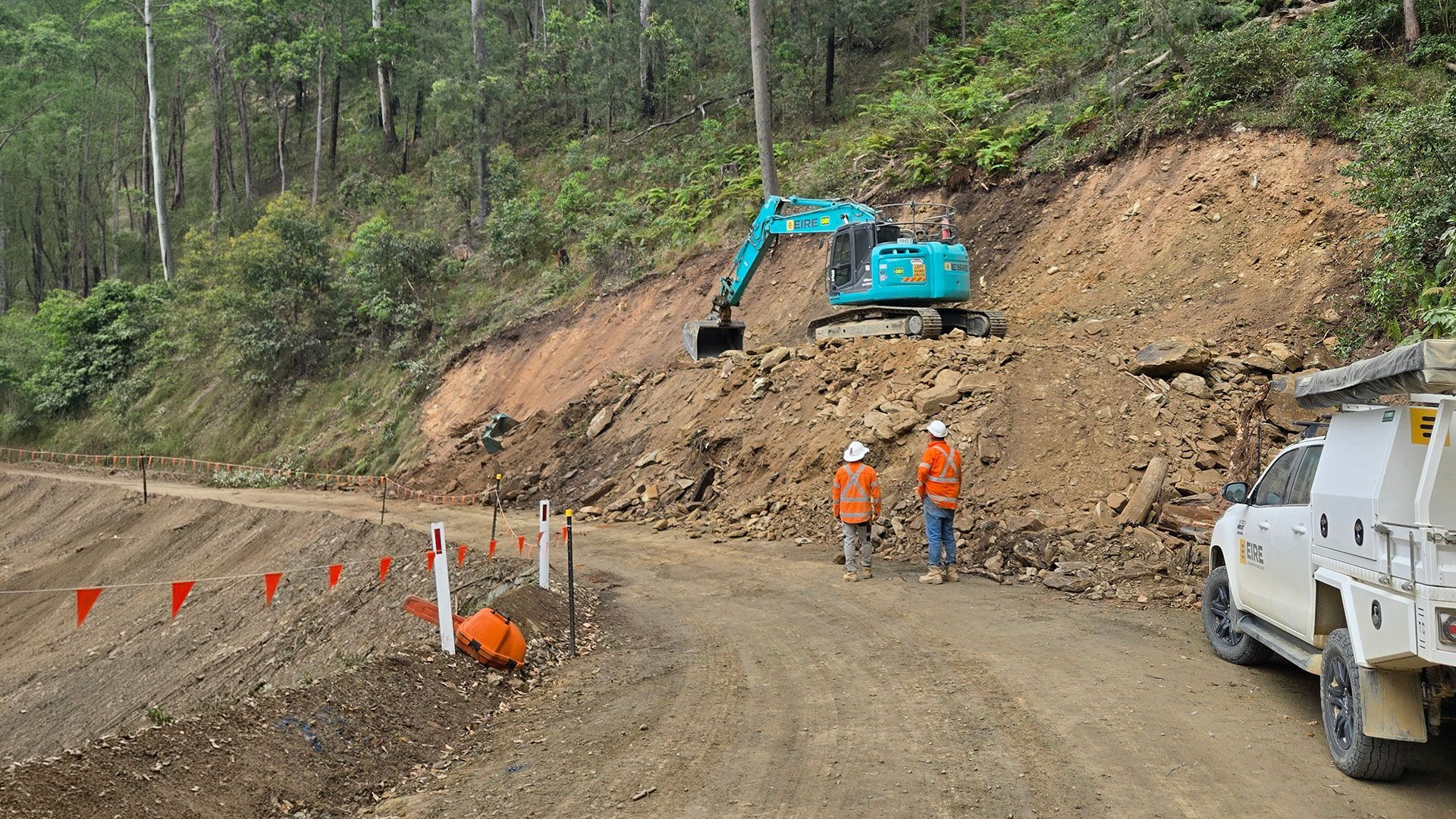 Construction crew with excavator clearing a dirt slope beside a muddy road.