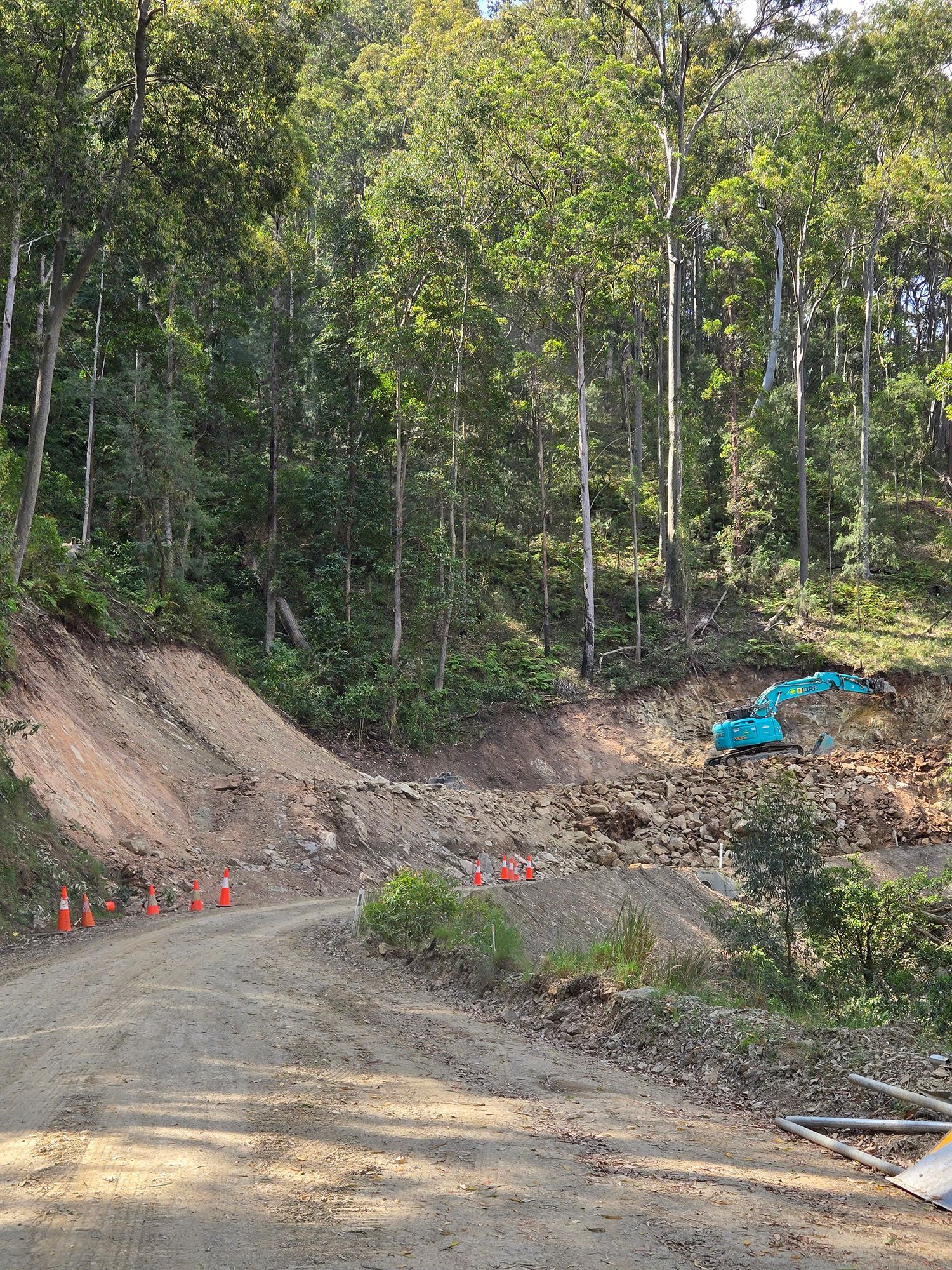 Gravel road under construction beside a dirt embankment in a wooded area, with orange markers and a blue tarp