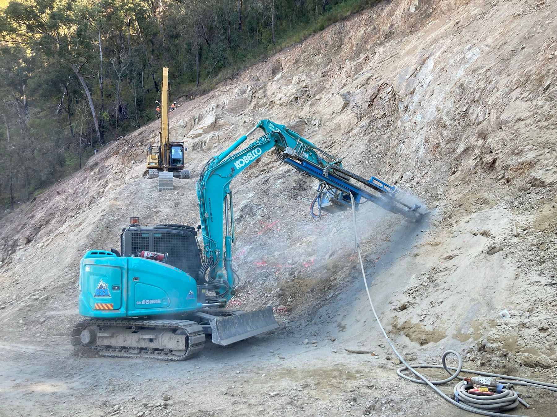 Blue excavator drilling into a rocky hillside, spraying dust at a construction site