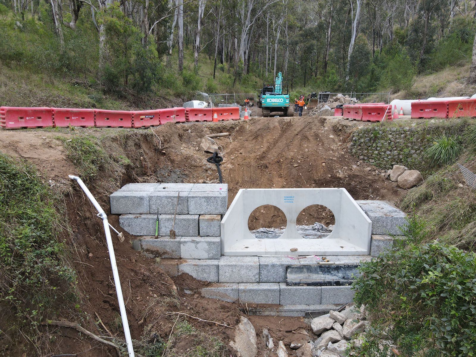 Excavation site with a concrete culvert and stone retaining wall under construction, red barriers around the pit.