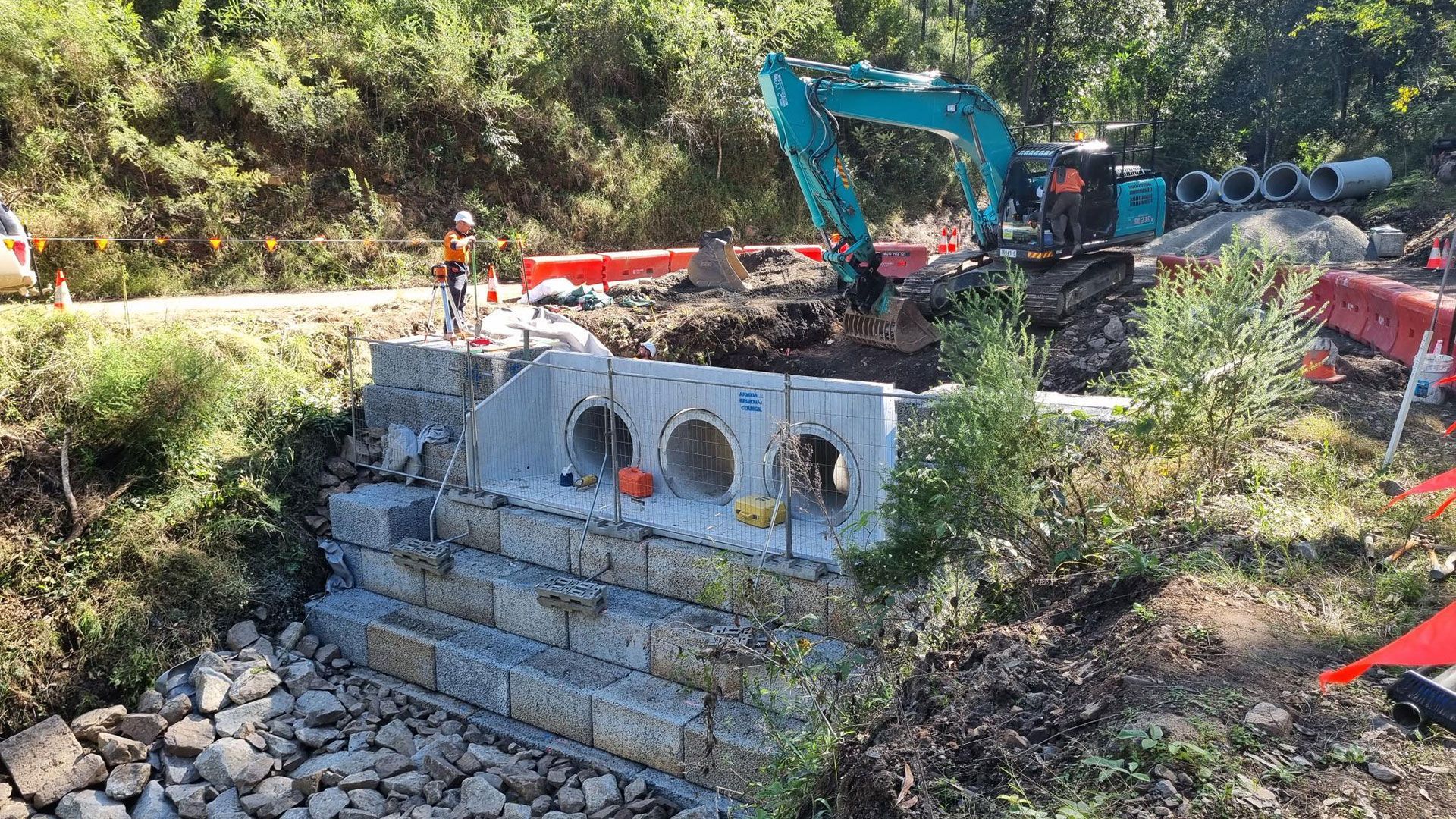 Construction site with excavator and stacked concrete culvert pipes beside a dirt road
