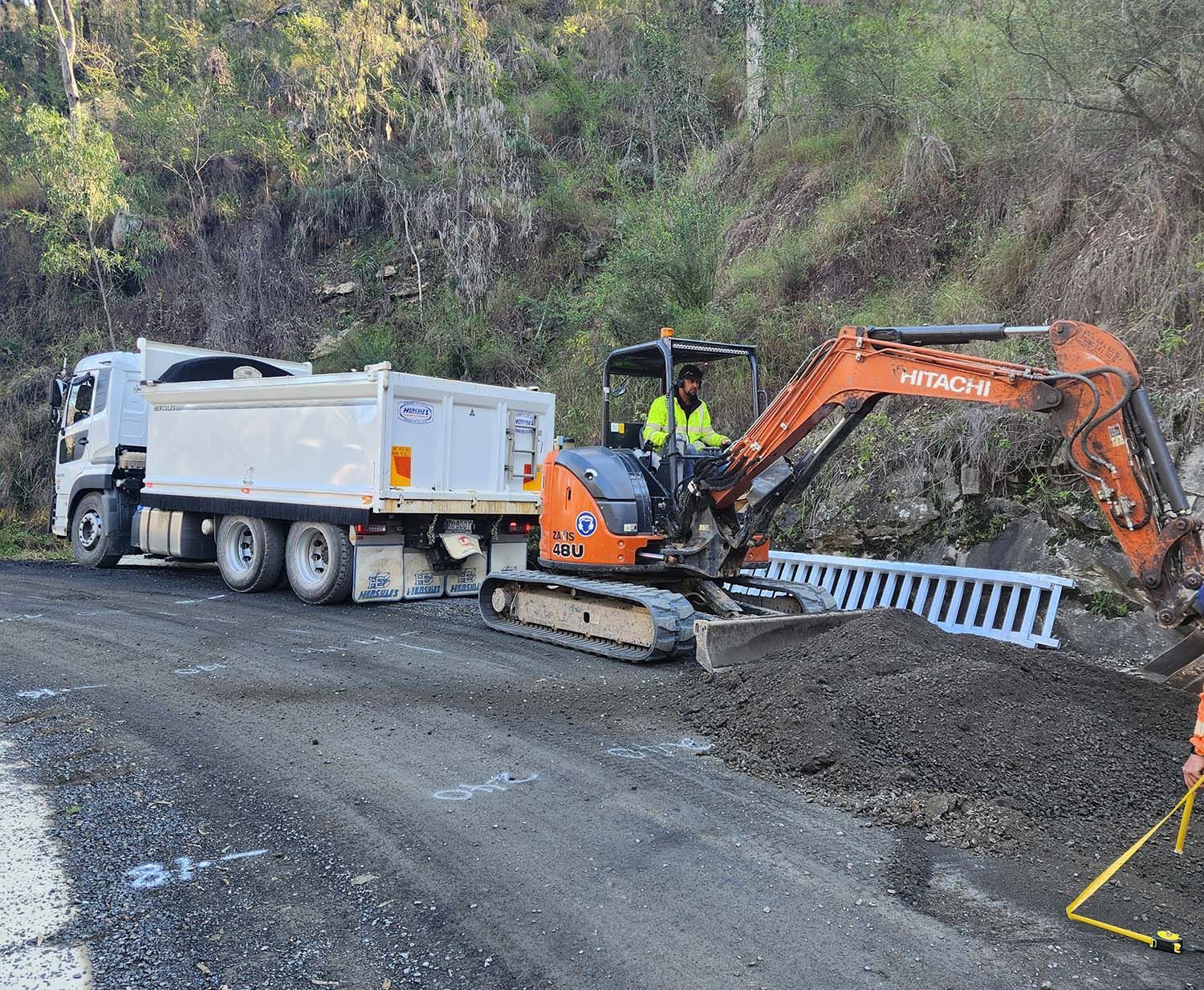 White dump truck and orange excavator working on a gravel road beside a hillside
