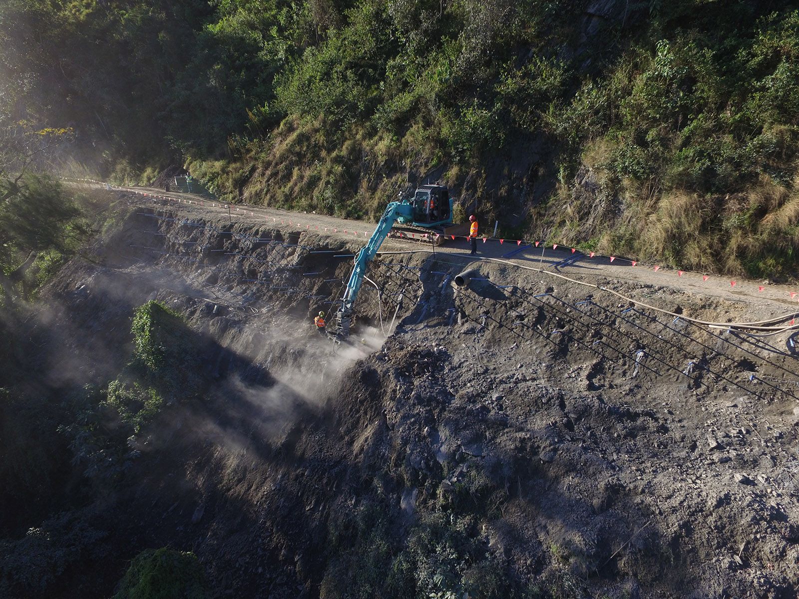 Excavator digging into a rocky hillside beside a dirt road, with workers nearby and dust rising.