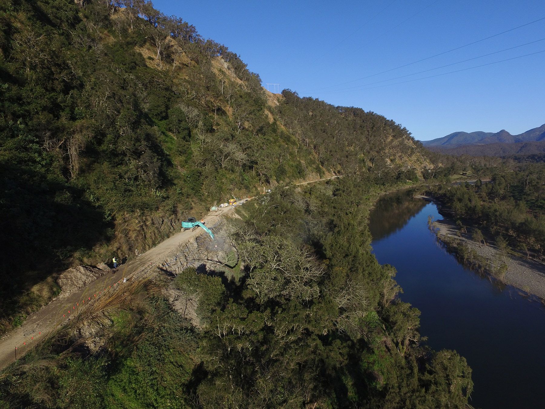 Narrow mountain road beside a river, with a small blue vehicle under a clear blue sky.
