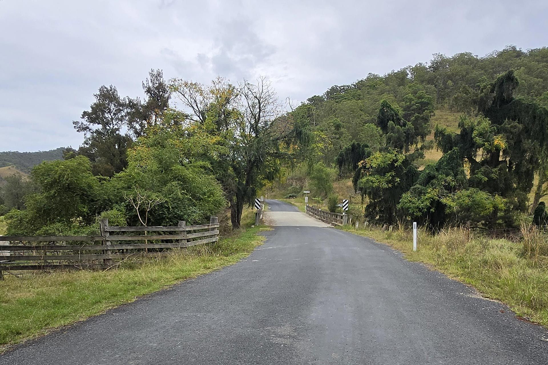 Gravel road curving through a green rural landscape with trees and wooden fencing under a cloudy sky