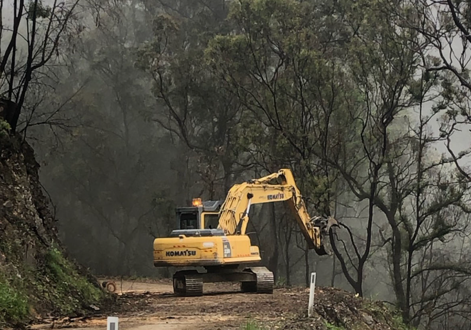 Yellow excavator on a dirt road amid smoky, burned forest trees