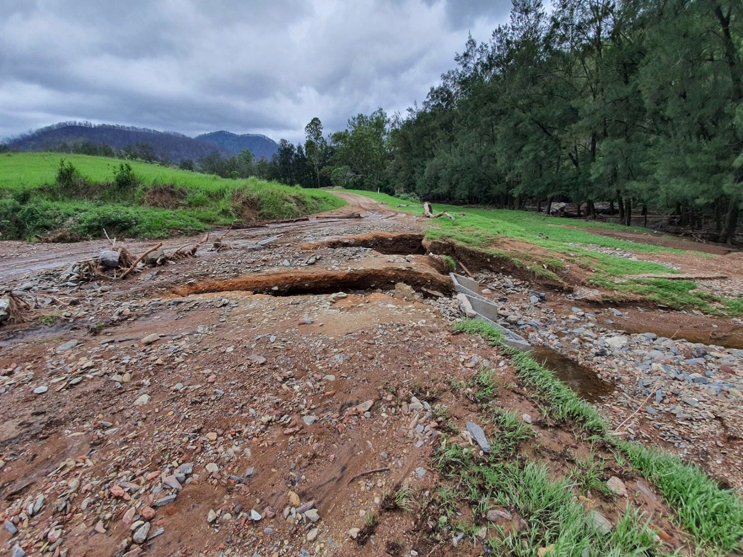 Broken dirt road washed out by erosion beside green fields and trees under a cloudy sky