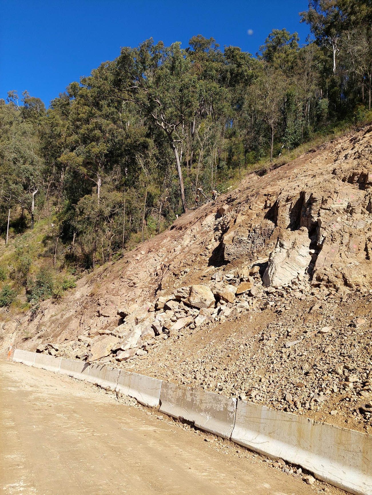 Rocky roadside slope with a concrete curb and trees under a blue sky