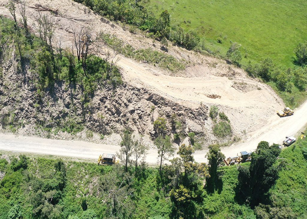 Dirt road along a green hillside with small construction vehicles and a cleared slope area.