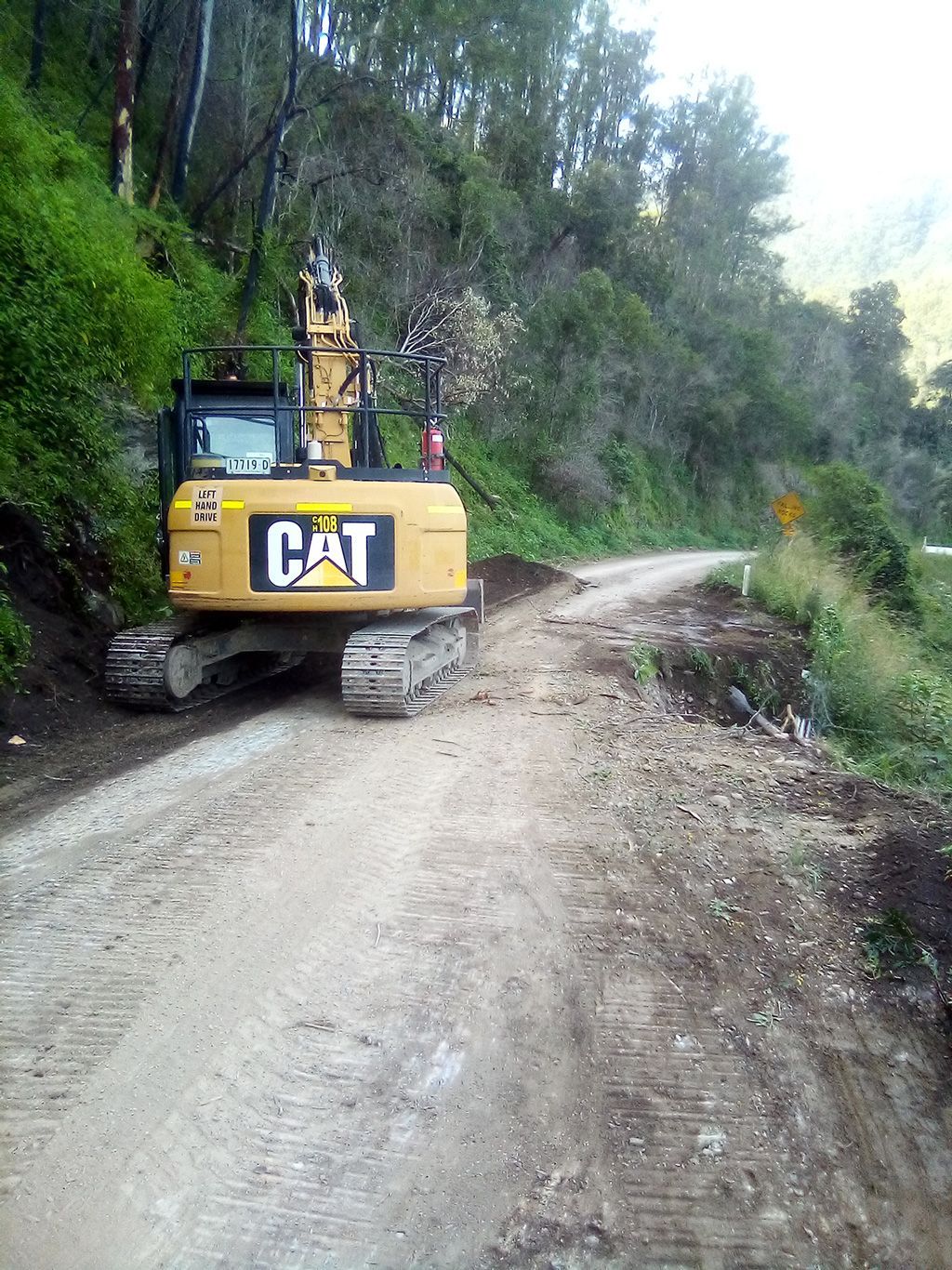 Yellow CAT excavator parked on a dirt road beside a wooded hillside.