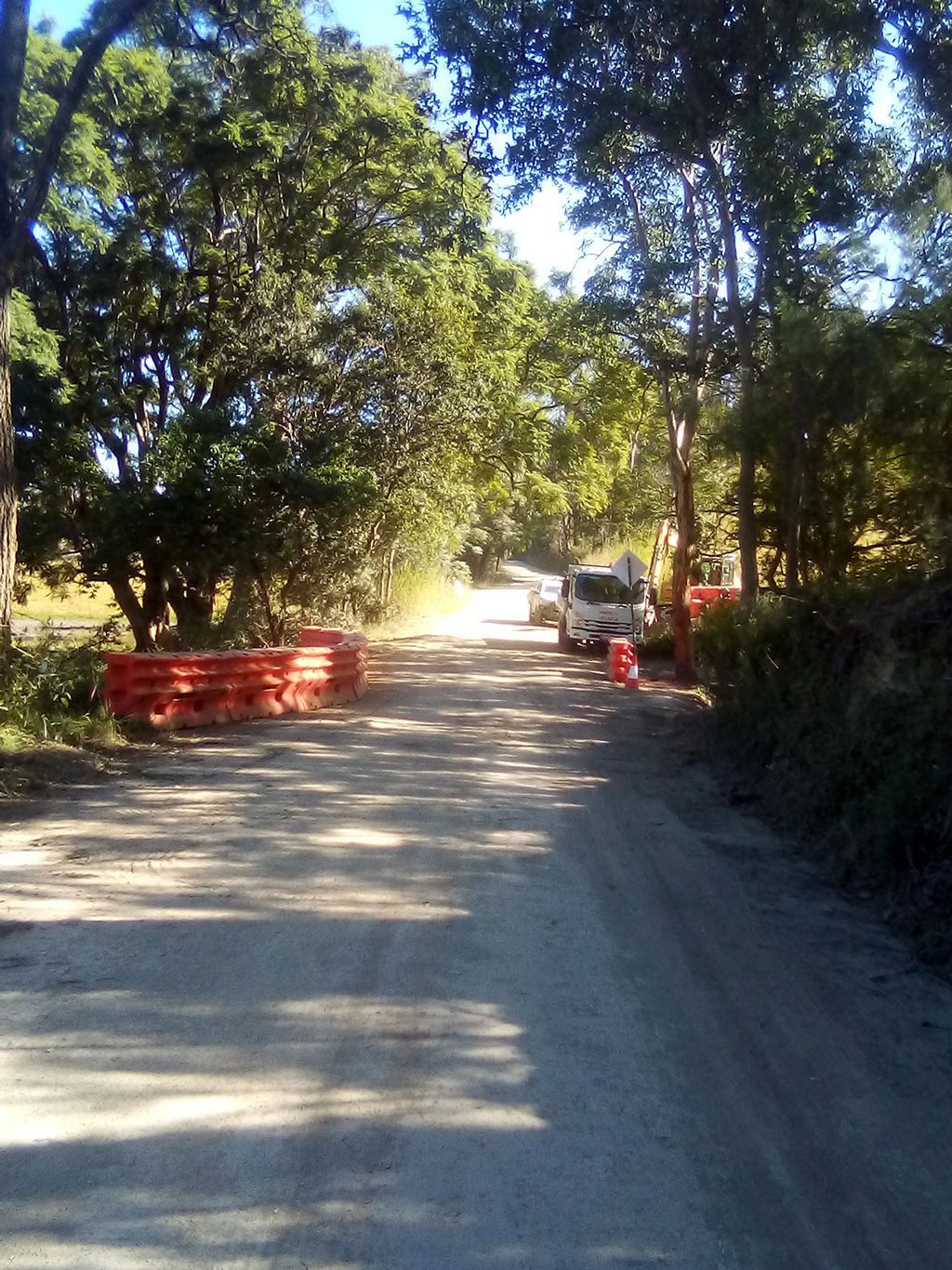Dirt road lined with trees, red barriers, and a parked vehicle in bright sunlight