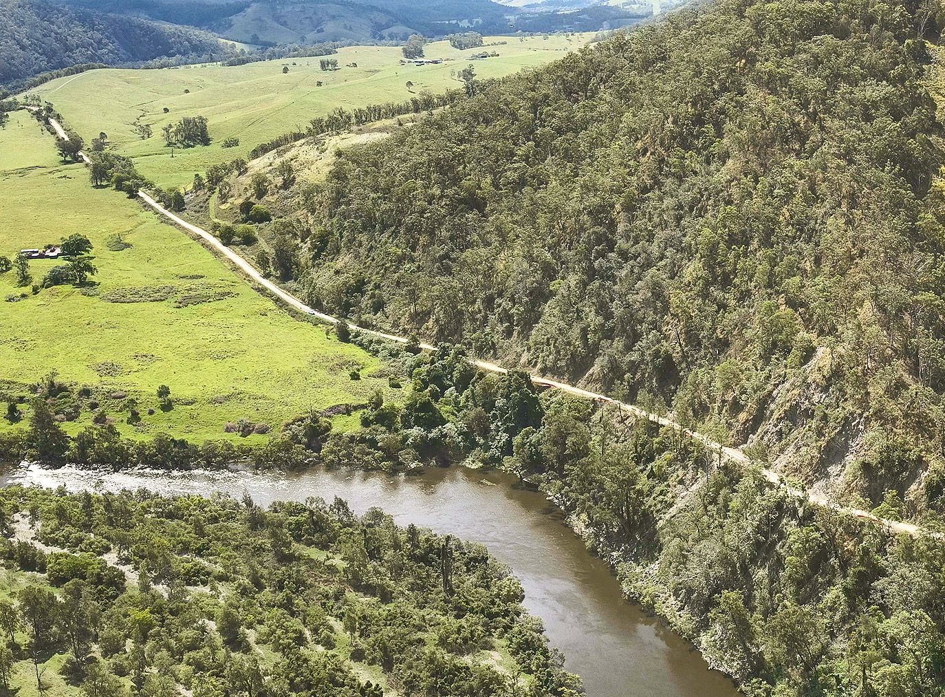 A winding river beside a steep green hillside and a narrow road through a lush valley.