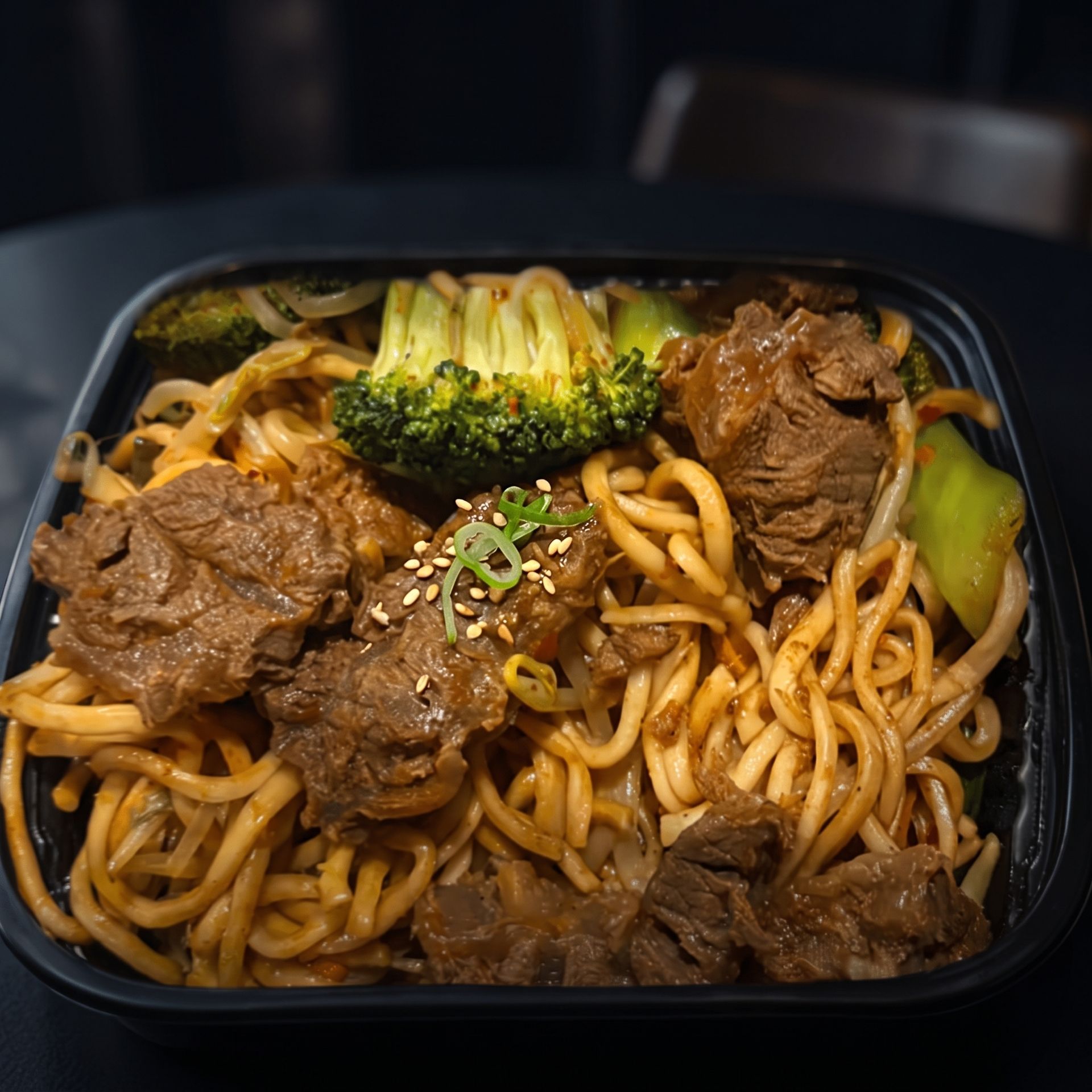 Black takeout container with stir-fried noodles, beef, broccoli, and green onions
