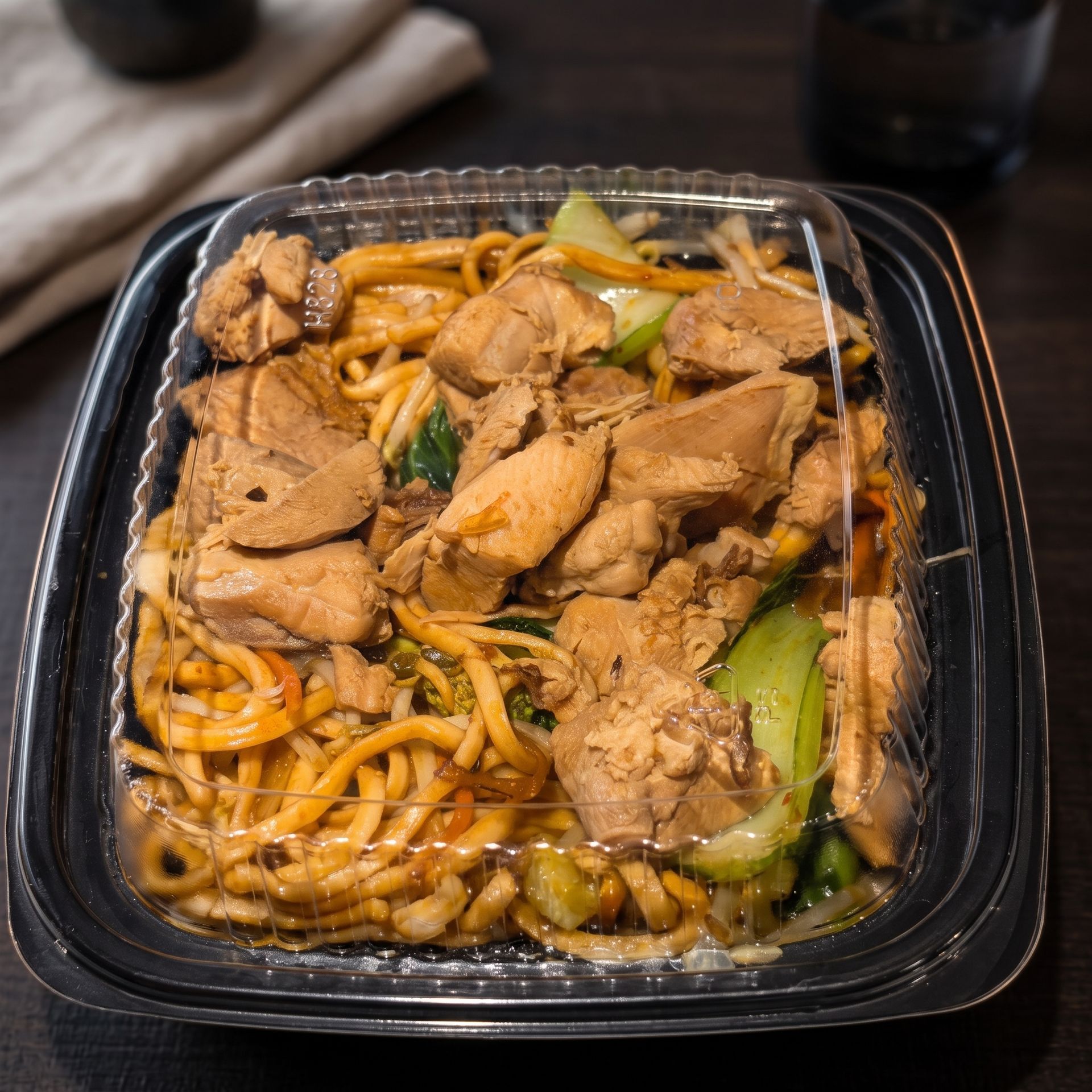 Takeout container with noodles, sliced chicken, and vegetables in brown sauce