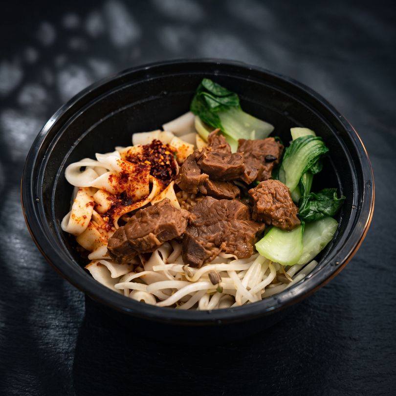Bowl of noodles topped with beef, bok choy, and chili oil on a dark surface