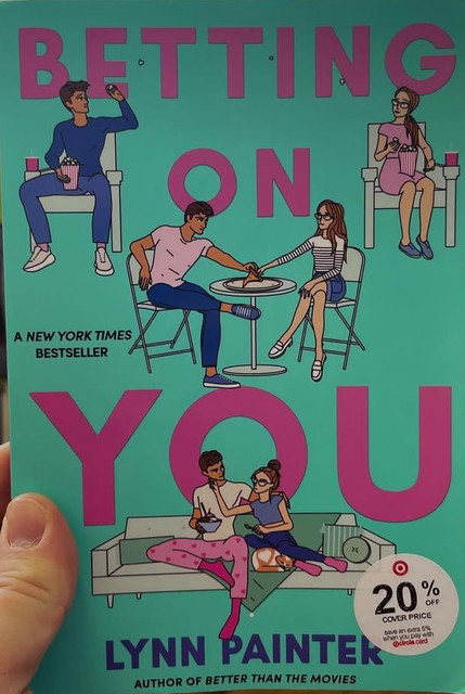A hand holds the book "Betting on You" by Lynn Painter, featuring a teal cover with illustrations of people.