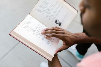 A person in a pink shirt holds an open book, their fingers resting on the page as they read.