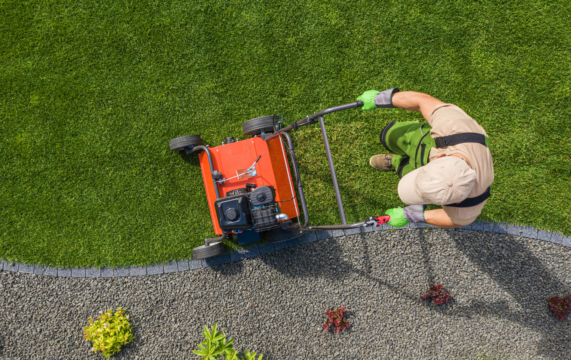 Person mowing a lawn beside a hedge, using an orange push mower on a gravel edge.
