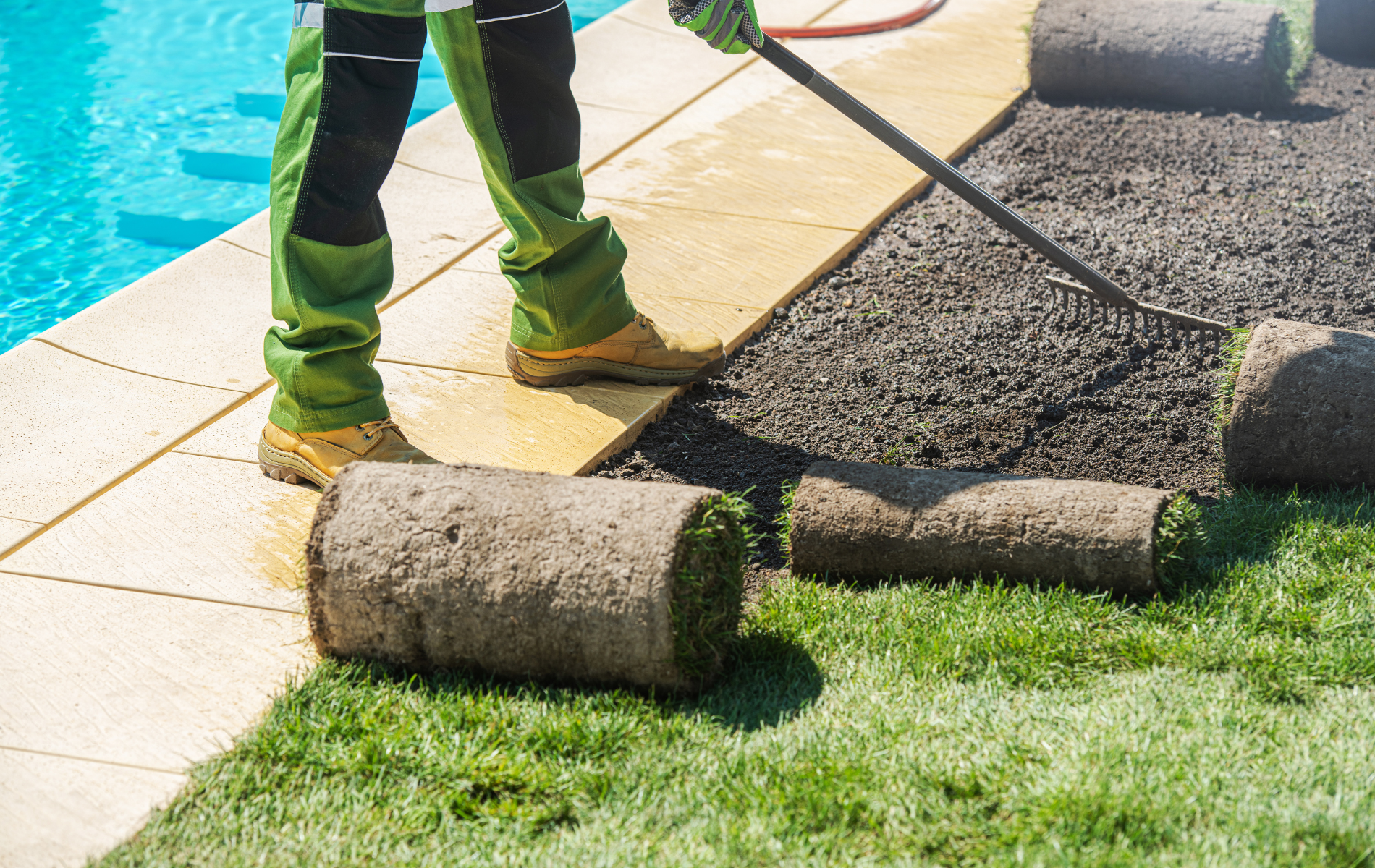 Person laying roll-out lawn beside a swimming pool, with grass and paving stones visible