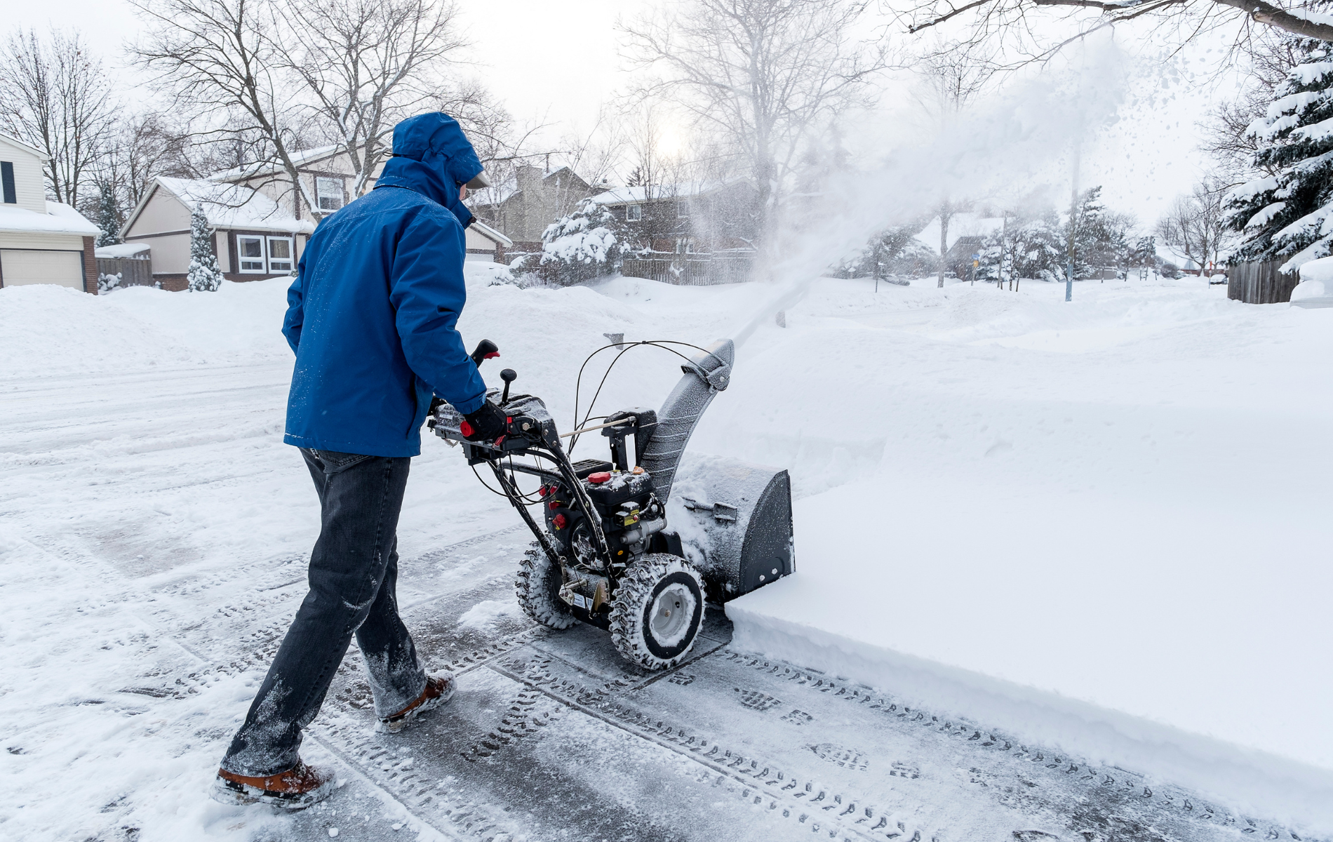 Person in a blue jacket shoveling snow with a snowblower on a snowy driveway