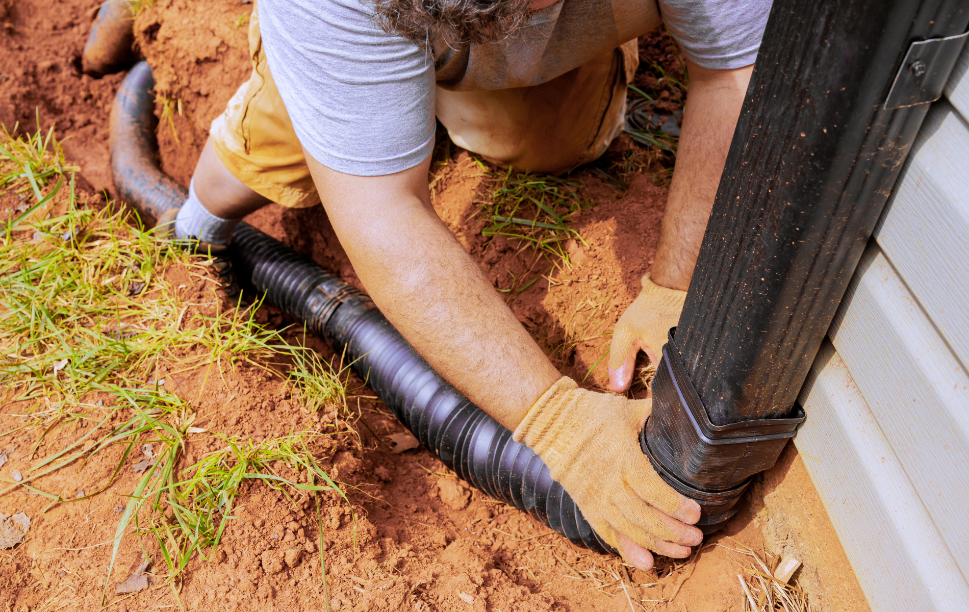 Person installing a black drainage pipe beside a house foundation in a dirt trench