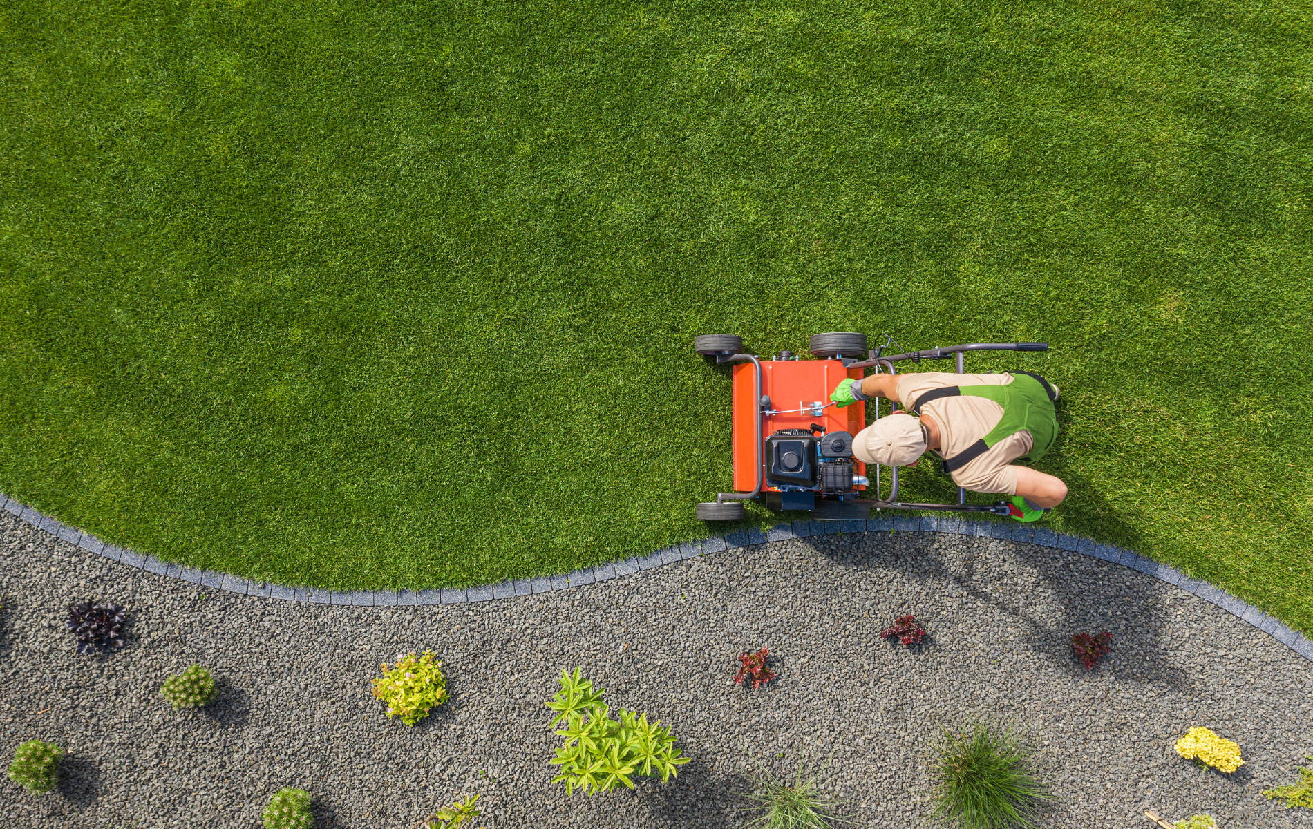 Worker using a lawn mower along the edge of a green lawn and gravel garden bed from above