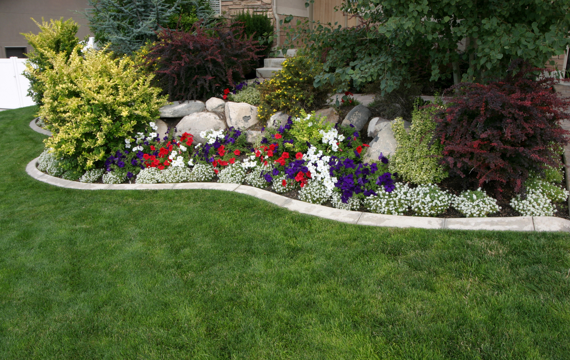 Landscaped garden bed with colorful flowers, shrubs, rocks, and a green lawn border