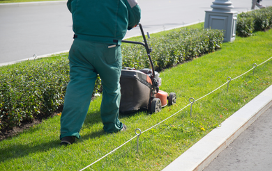 Worker mowing grass along a roadside sidewalk with a push mower and string trimmer