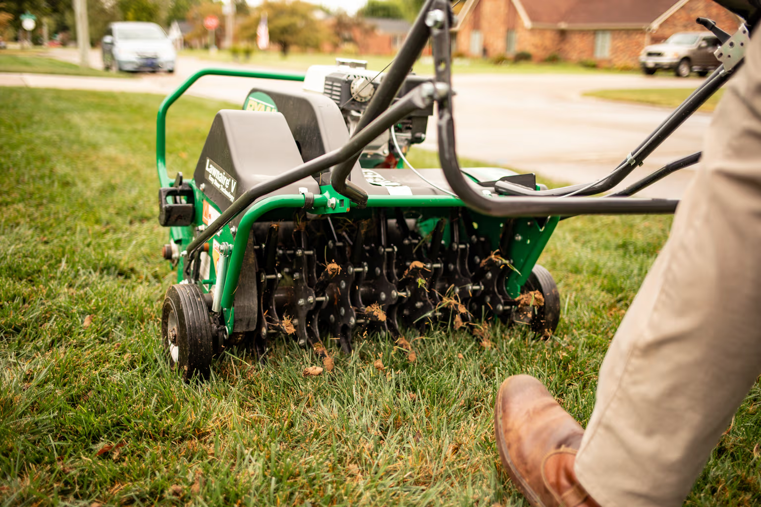 Person using a green lawn aerator on a grassy yard near a sidewalk