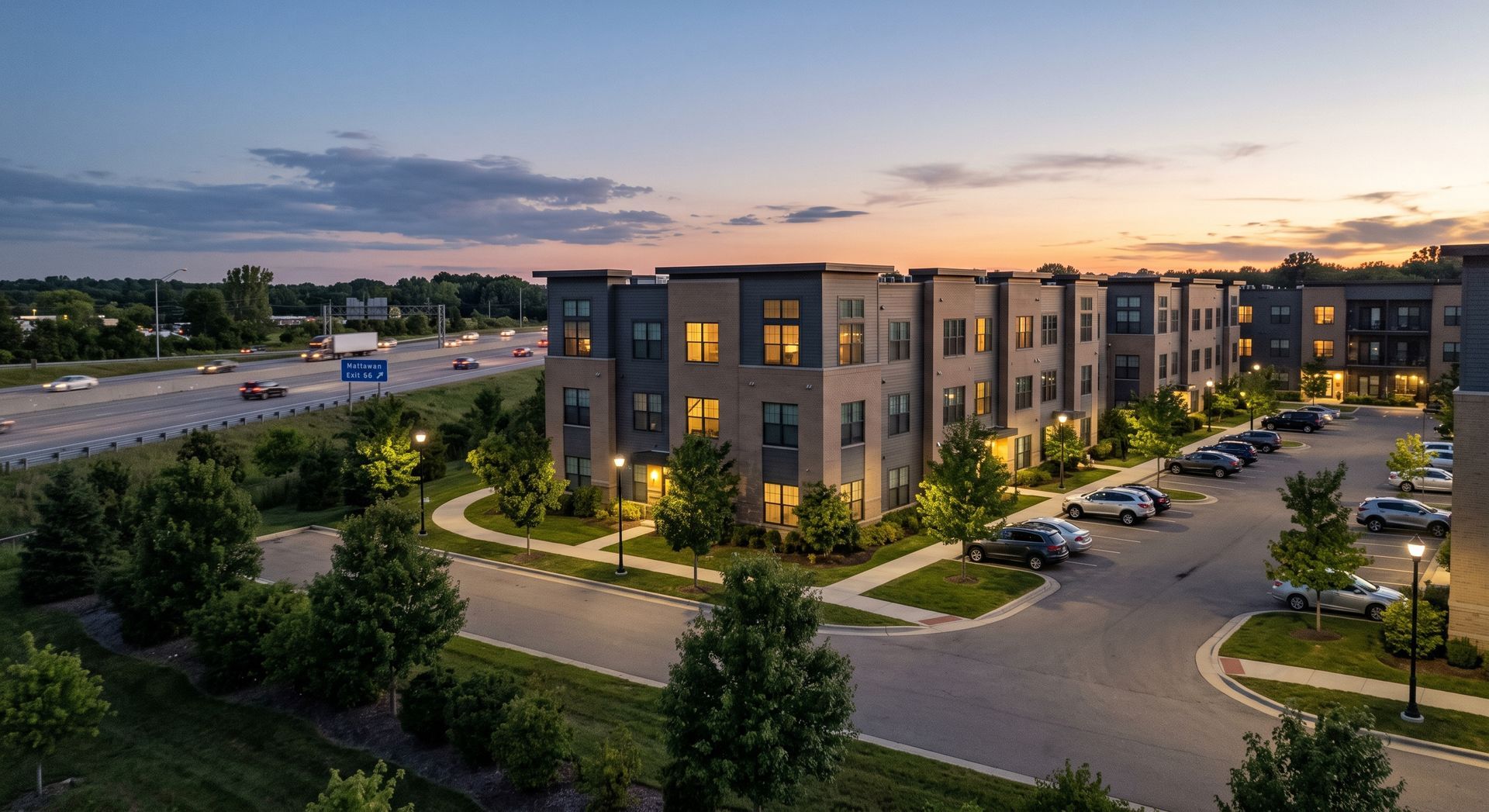Modern apartment complex at dusk with lit windows, landscaped paths, and parked cars