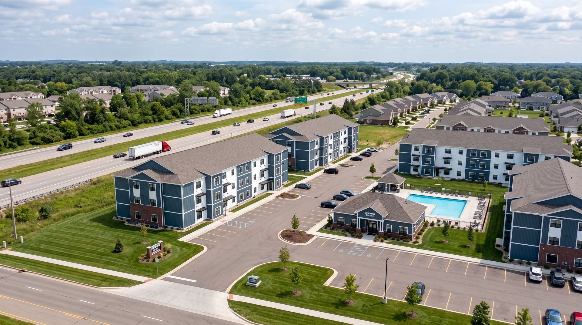 Aerial view of modern apartment complex with parking lots, pool, and nearby highway.