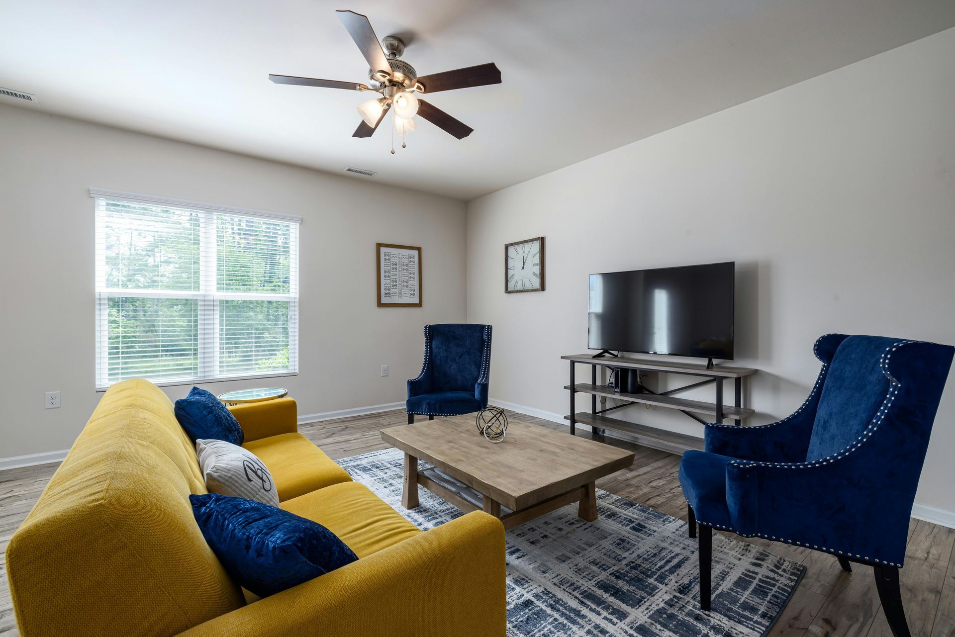 Bright living room with yellow sofa, blue chairs, coffee table, TV, ceiling fan, and window light