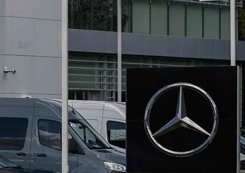 Mercedes-Benz dealership sign outside a glass-front building with parked vans nearby