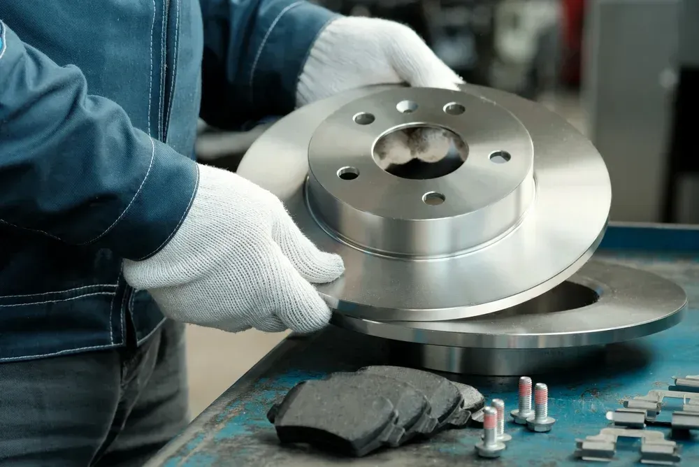 Gloved worker handling a silver brake disc on a workshop table with tools and parts nearby