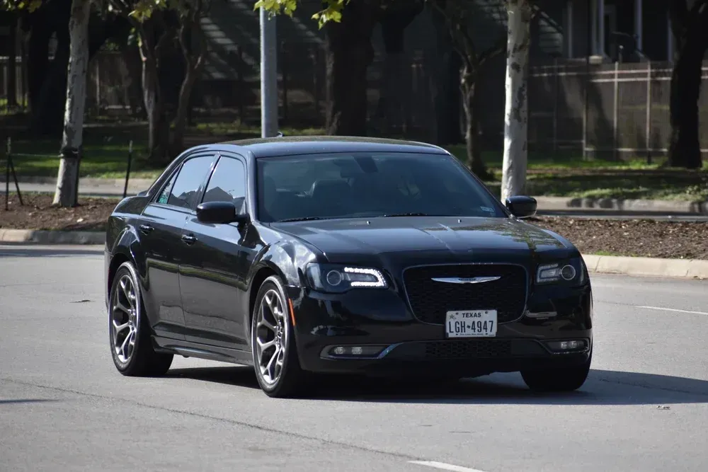 Black sedan driving on a road beside trees and a fence
