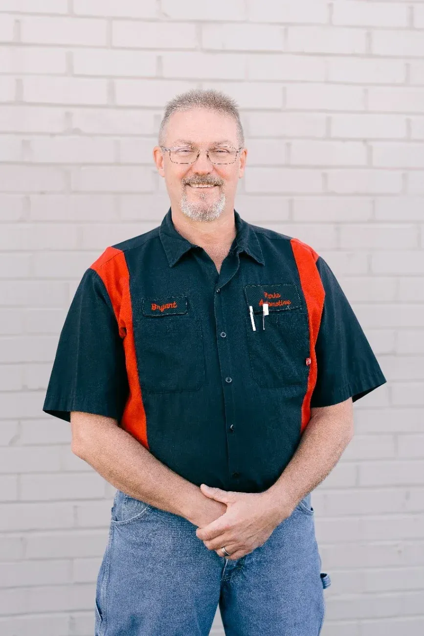 Man standing against a white brick wall, wearing a dark shirt with red suspenders and blue jeans.