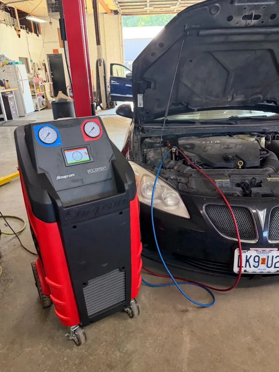 Red automotive AC recharge machine beside a car with its hood open in a garage