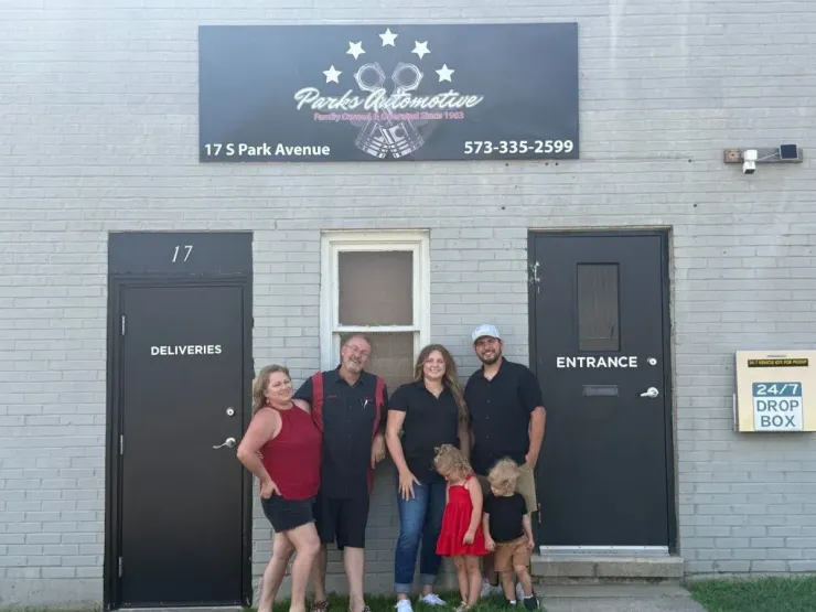 Four people standing outside a gray building with black doors and a sign above the entrance