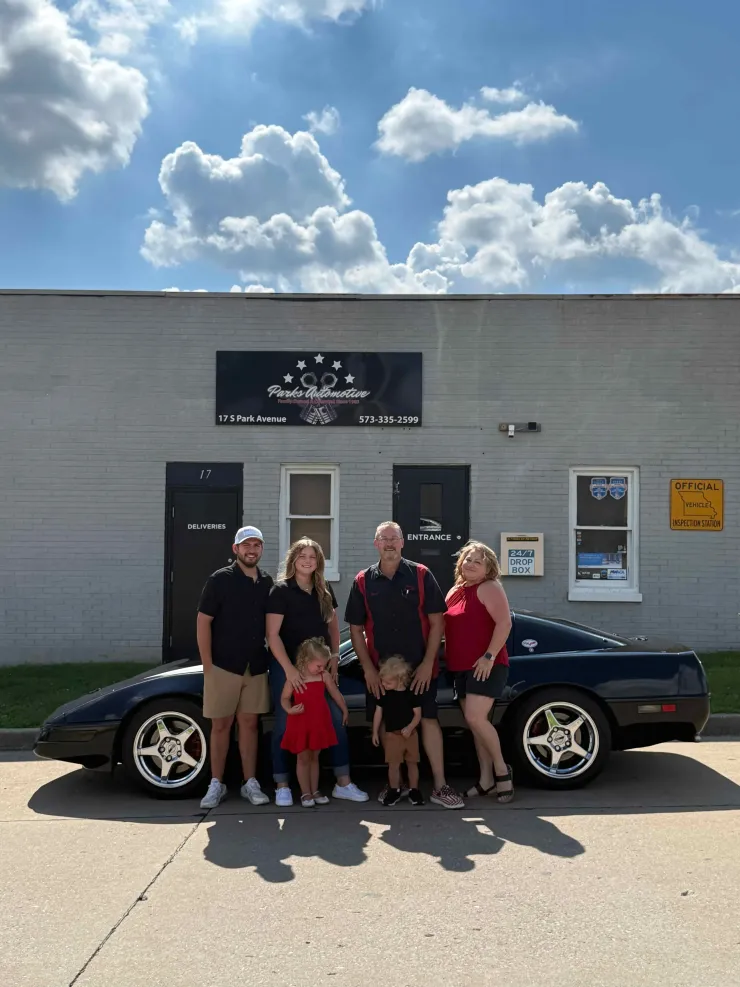 Five people posing in front of a black car outside a building under a cloudy sky.