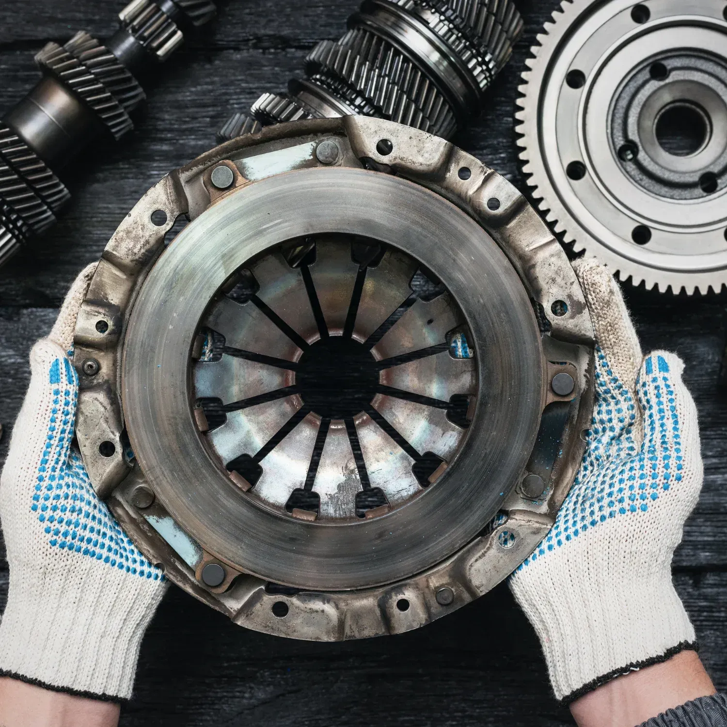 Gloved hands holding a metal clutch assembly with gears and mechanical parts on a workbench