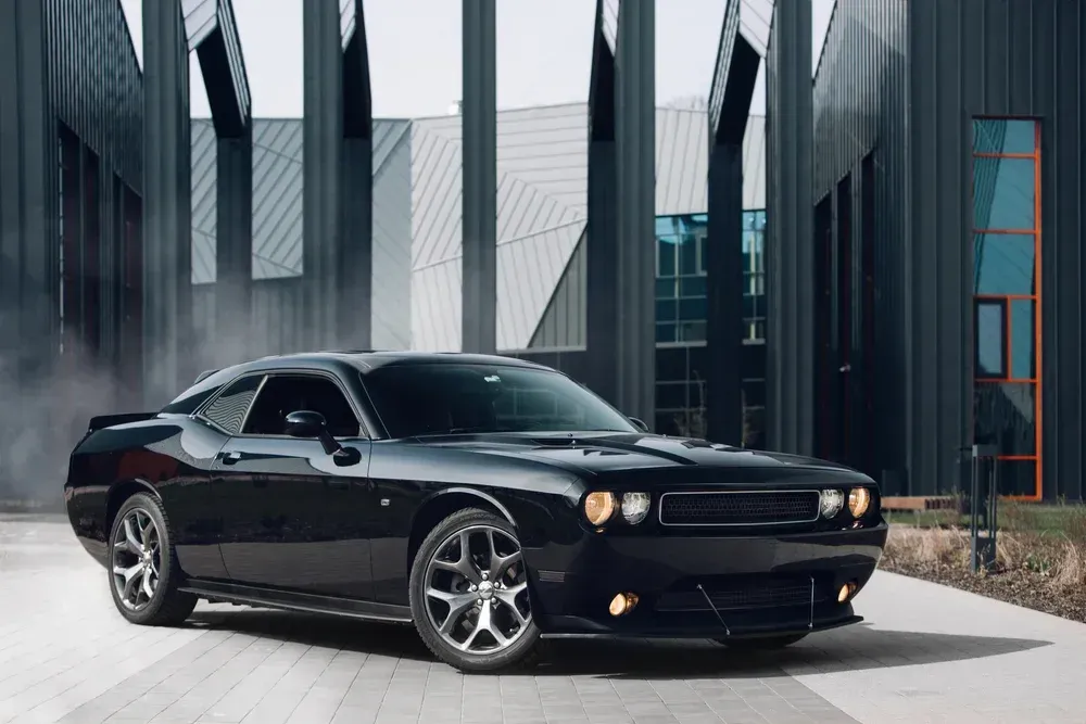 Black classic muscle car parked beside modern glass buildings.