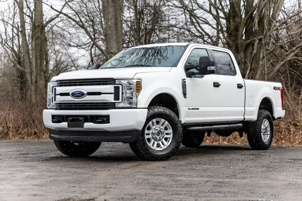 White Ford Super Duty pickup truck parked on a wet gravel road beside leafless trees