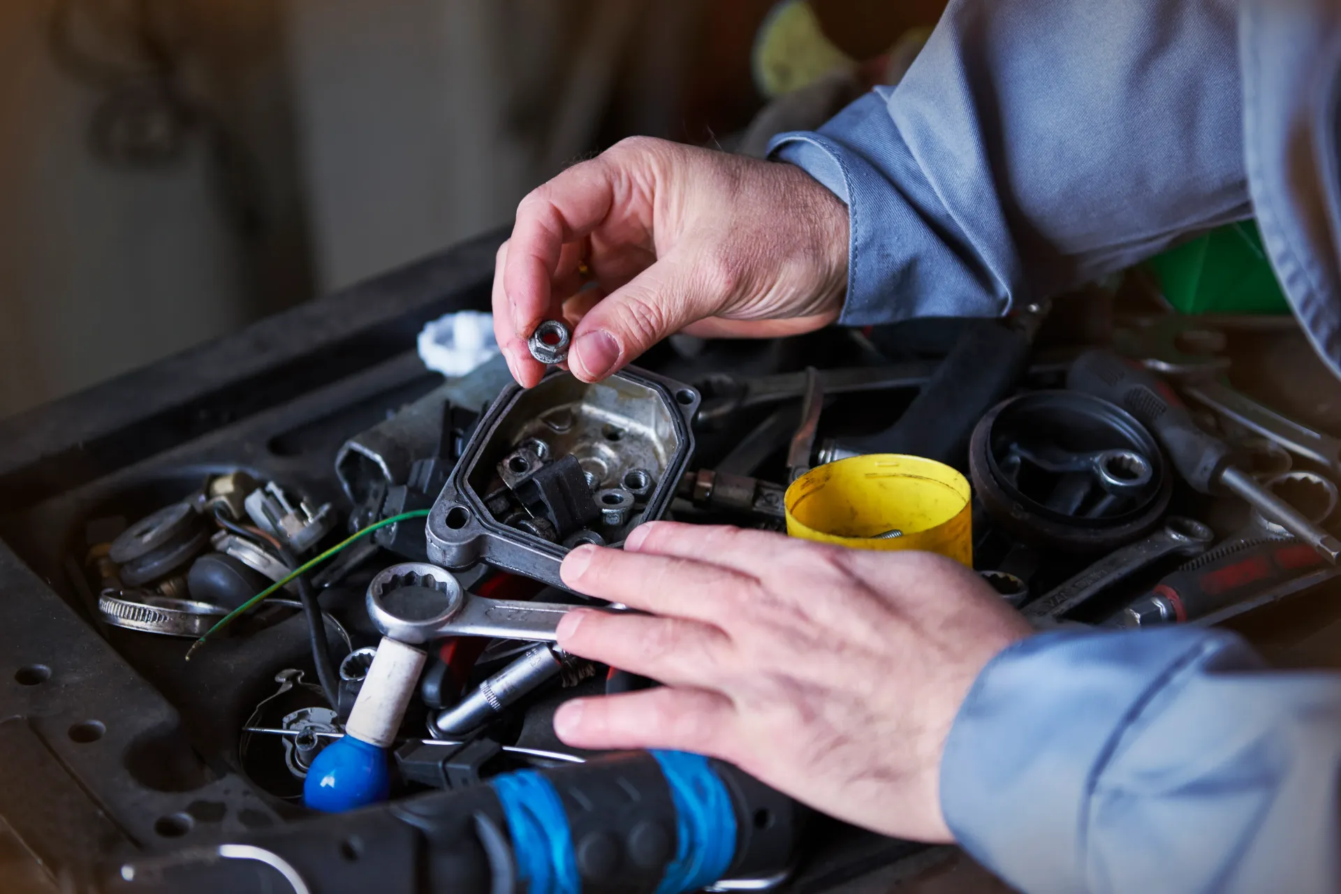 Hands working over a cluttered repair bench with tools, wires, and a yellow container