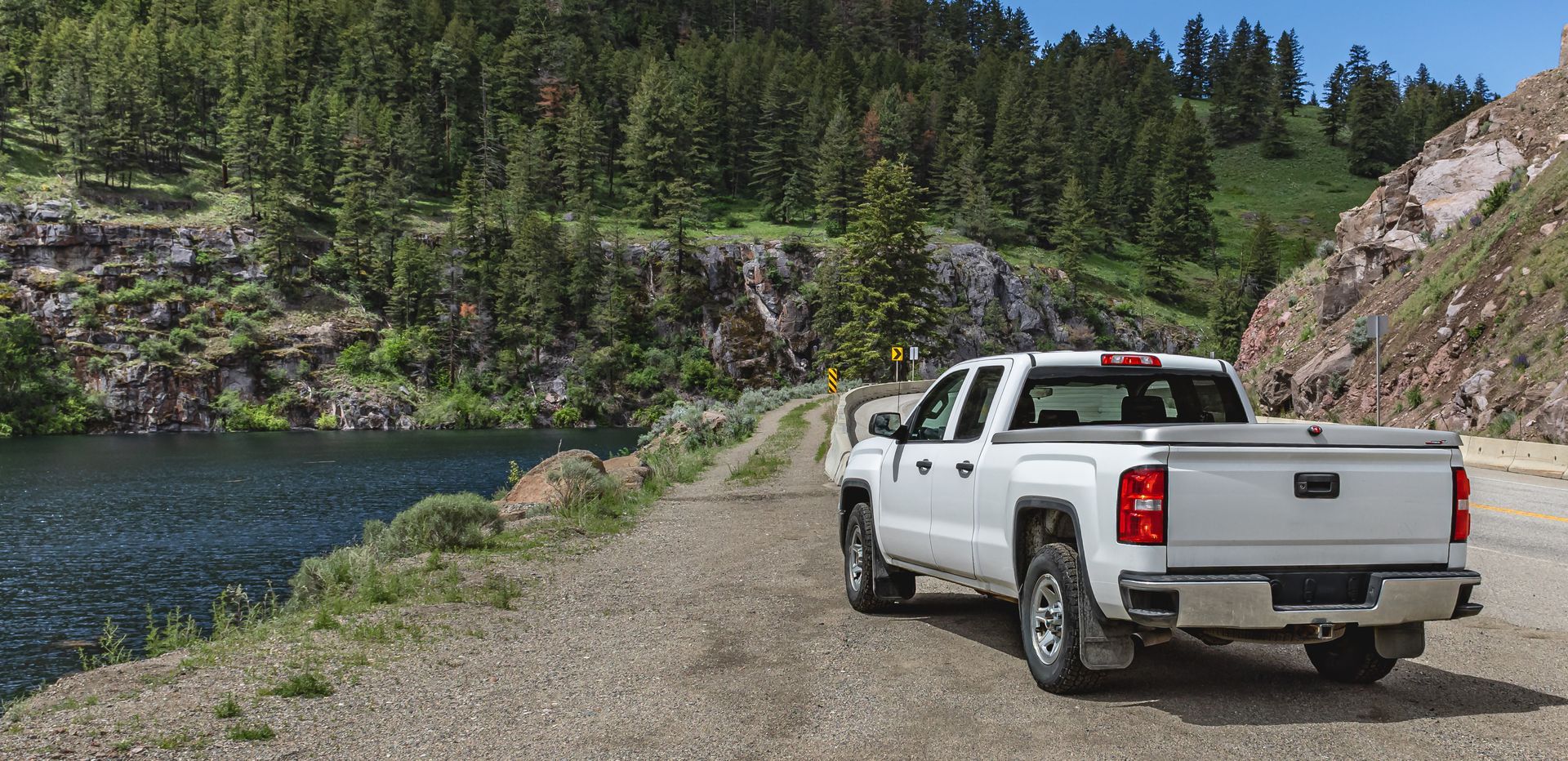 White pickup truck parked on a gravel road beside a river and forested hillside under a blue sky