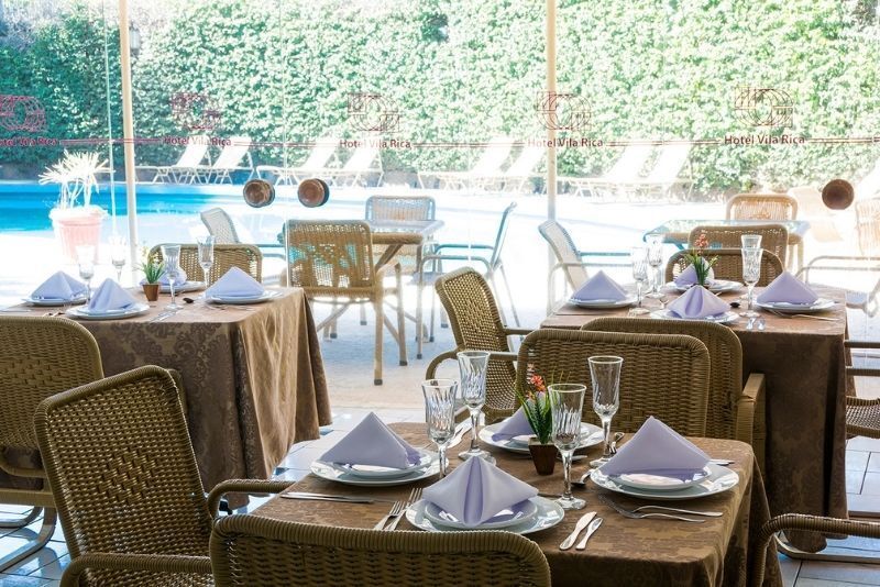 Pátio externo do restaurante com cadeiras de vime e mesas postas com vista para uma piscina iluminada e área verde.