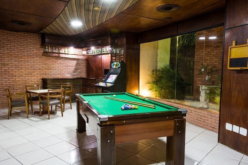 Sala de bilhar aconchegante com mesa de sinuca verde, paredes de tijolos, piso de azulejo e máquina de jogos eletrônicos.