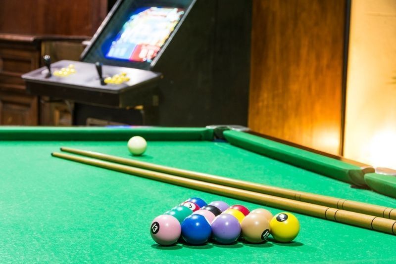 Mesa de bilhar com bolas enfileiradas e um taco, em uma sala de jogos com pouca luz.