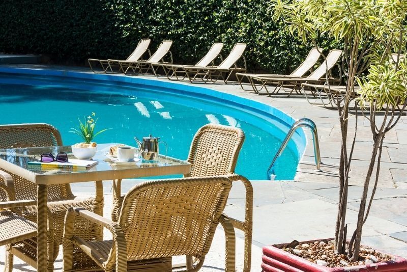 Pátio à beira da piscina com cadeiras e mesa de vime ao lado de uma piscina azul, rodeado por espreguiçadeiras e árvores.