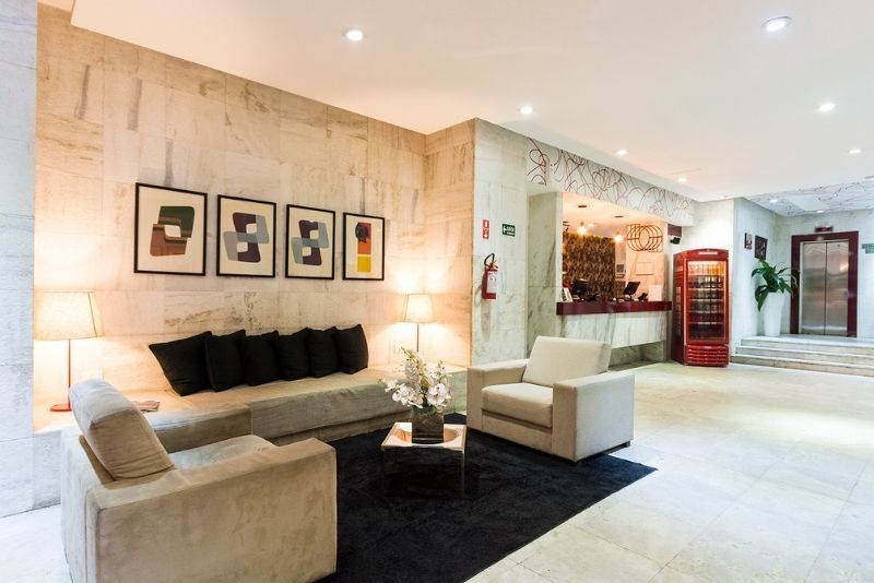 Hall de entrada de hotel moderno com sofás bege, tapete preto, obras de arte emolduradas e decoração com detalhes em vermelho.