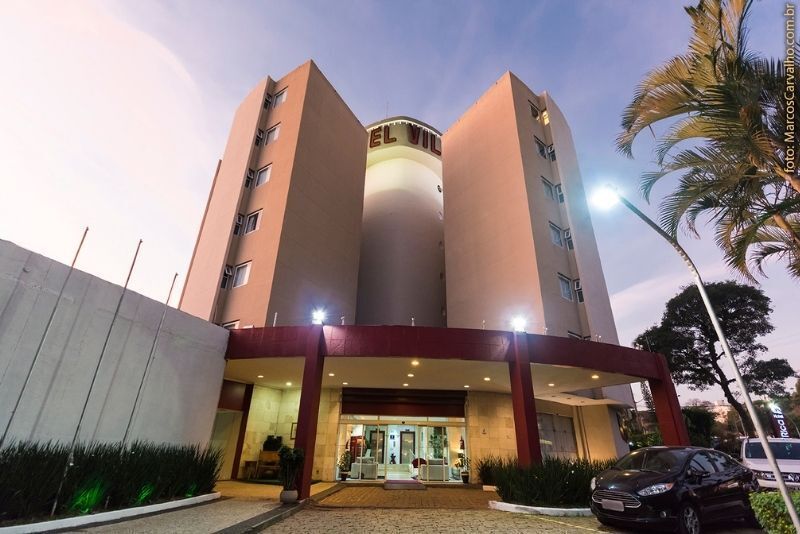 Entrada de hotel moderno ao entardecer, com fachada alta e curva, saguão iluminado, palmeiras e carro estacionado.